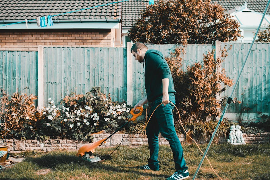 A man wearing glasses, a dark hoodie, and jeans, is mowing a lush green lawn with a bright orange electric grass trimmer in a residential garden. The garden features a flower bed with blooming white a