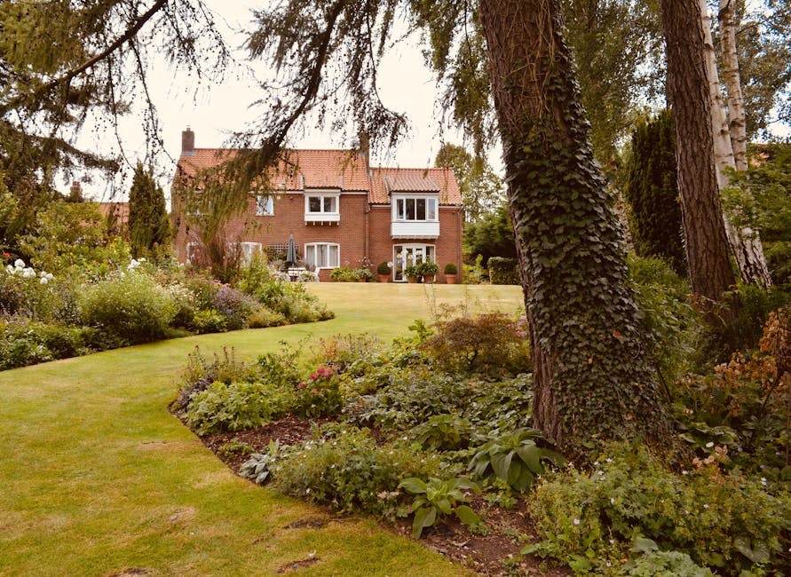 A spacious garden showcasing well-maintained lawn areas bordered by lush flower beds with various flowering plants and greenery, framed by tall trees including one with a textured, ivy-covered trunk. 